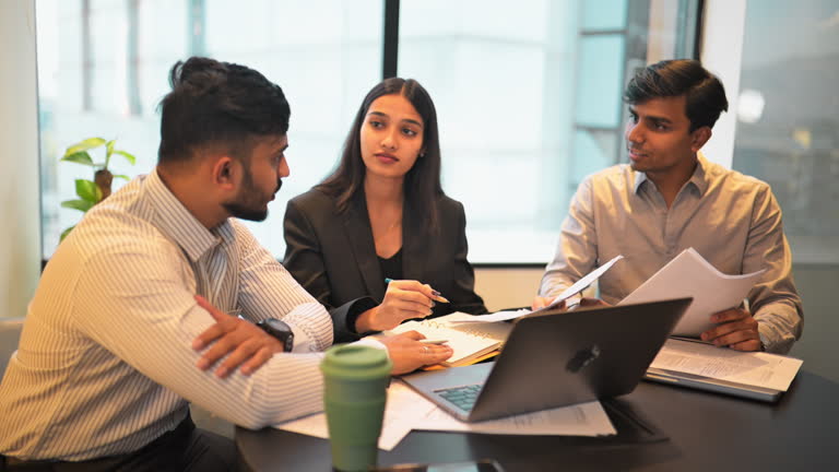 Young male and female adults discussing in a modern meeting room. The team is seated around a conference table, reviewing papers and discussing work, with a laptop open in front of them. The setting exudes a professional yet dynamic atmosphere, with the colleagues engaging in productive conversation, sharing ideas, and collaborating on their project